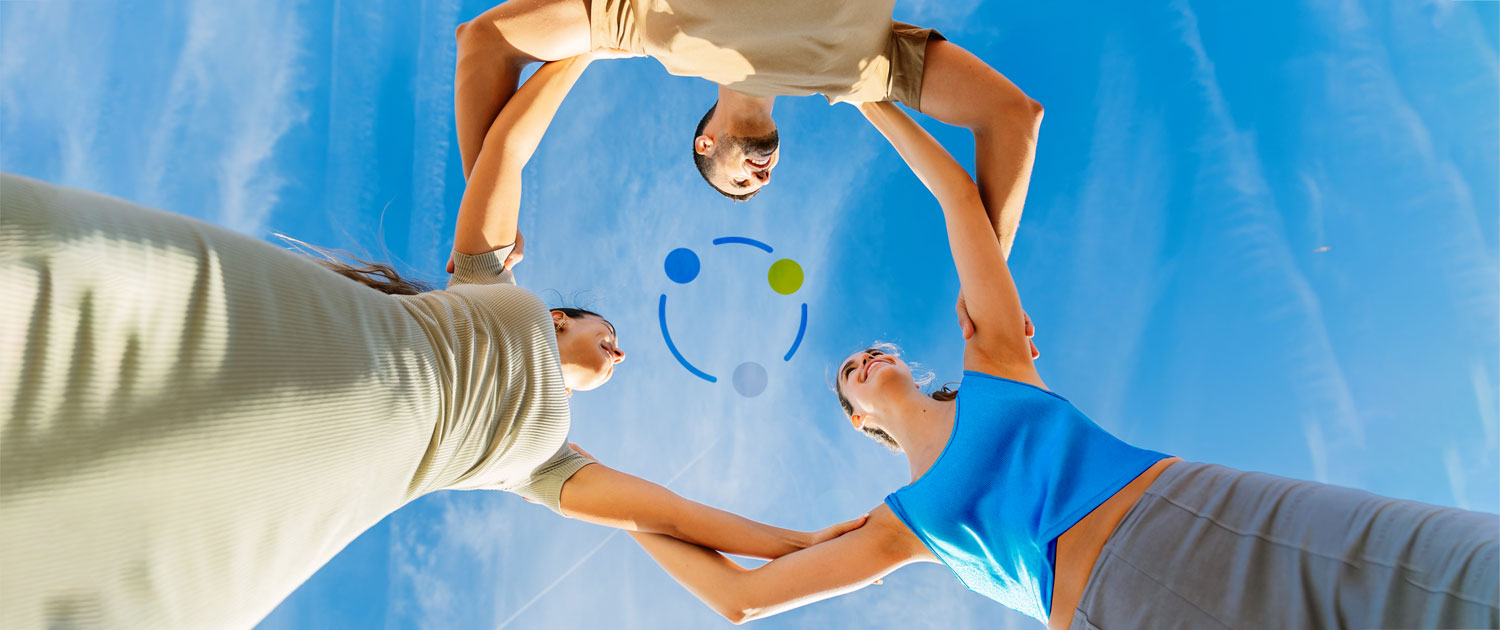 A young man and two young women are photographed from the ground, looking upwards. They stand in a circle, holding each other's shoulders. Above them, the blue sky with a few white clouds. The three are smiling at each other. In the center of the image, three dots with blue lines also form a circle.