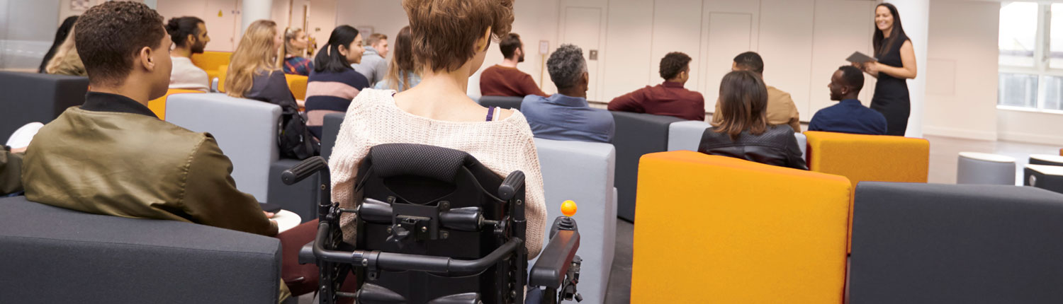 A young female speaker in an elegant black dress stands facing the audience in front of four rows of comfortable, upholstered armchairs. Around a dozen people are seated in the armchairs, they are displayed from behind. In the last row, in the center of the image, a person in a wheelchair is amongst them.