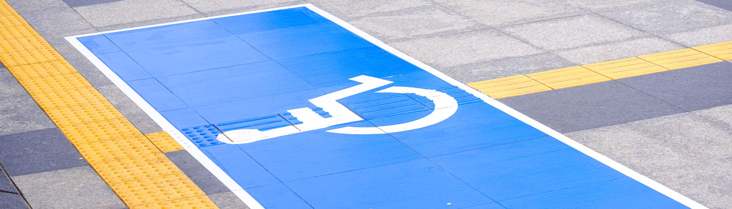 On a sidewalk paved in two shades of gray, yellow tactile paving strips guide people with visual impairments. In the center of the image, a larger rectangular area in bright blue with a white border with a pictogram of a person in wheelchair in white.