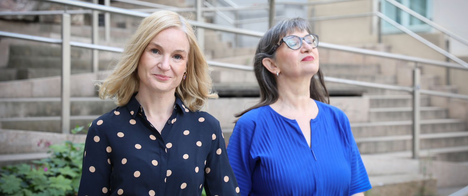 Two women in close-up. They are sitting side by side. The woman in the foreground is smiling at the camera. She is blonde and wearing a blue top with beige polka dots. It is Judith Platter. The woman in the background has her eyes closed and appears very relaxed. She has long, brown hair. She is wearing distinctive glasses with brown frames and a blue top. It is Ursula Nasswetter.