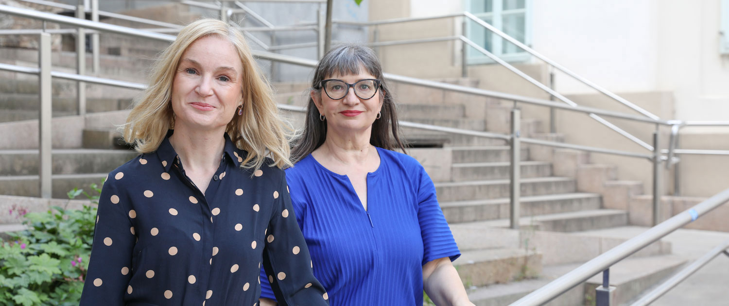 Two women in close-up. They sit side by side and smile at the camera. The woman in the foreground is blonde and wears a blue top with beige polka dots. It is Judith Platter. The woman in the background has long, brown hair. She wears distinctive glasses with brown frames and a blue top. It is Ursula Nasswetter.