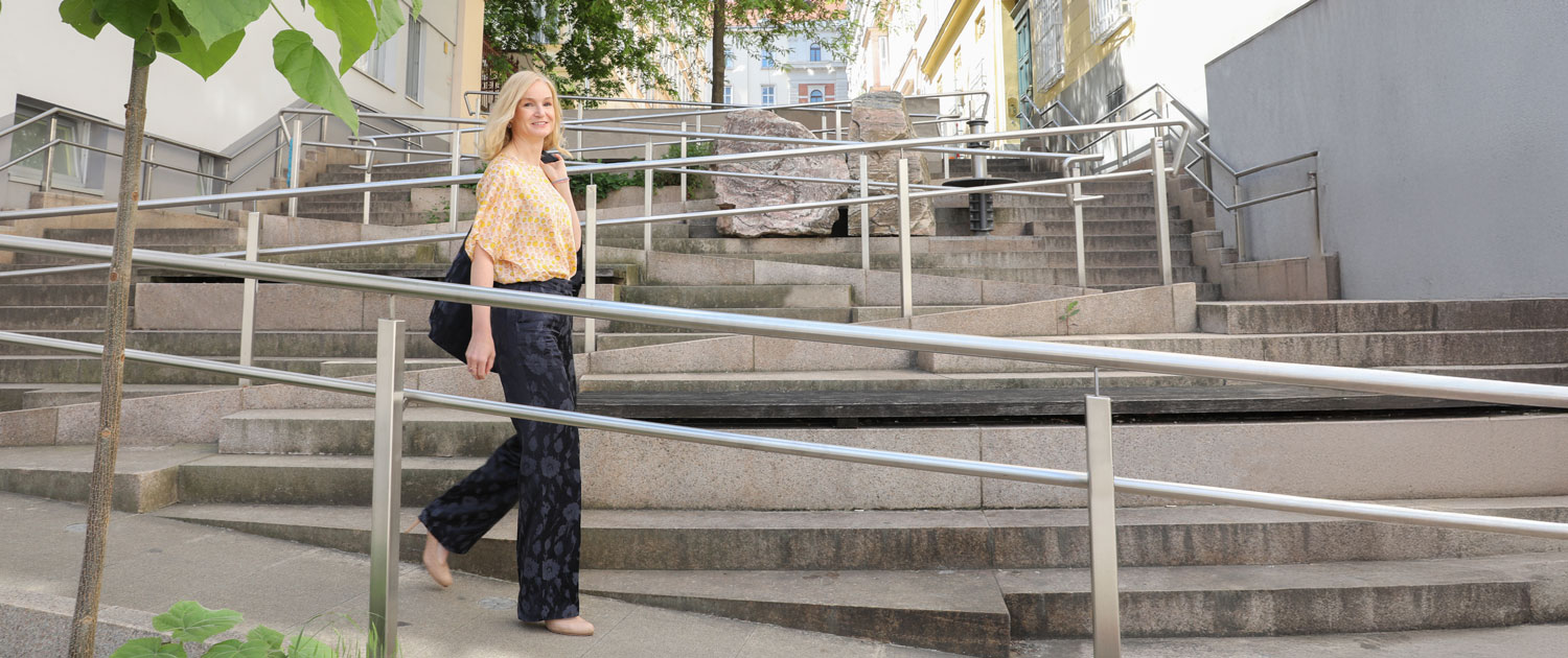 A middle-aged woman wearing a blue pantsuit with beige polka dots stands in the middle of a staircase. She smiles at the camera and leans against the metal railing of a ramp that runs up the stairs. It is Judith Platter. The staircase is the Thurnstiege, with accessible ramps in Vienna's 9th district.