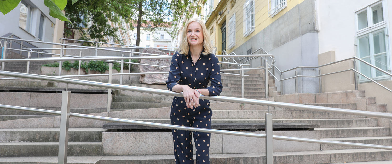 A middle-aged woman wearing a blue jumpsuit with beige polka dots standing on a staircase. She smiles at the camera and leans against the metal railing of a ramp that runs up the stairs. It is Judith Platter. The staircase is the Thurnstiege, with accessible ramps in Vienna's 9th district.