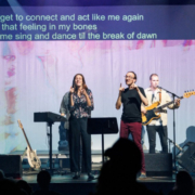 Five people are visible on a stage. They stand in front of a screen. At the top of the screen are three lines of the English lyrics to the song being sung. These lyrics are produced by sign language interpreters. The two people in the middle of the stage are sign language poets. They perform the text in sign language. © Beat the Silence with link: Das WUK | Beat the Silence