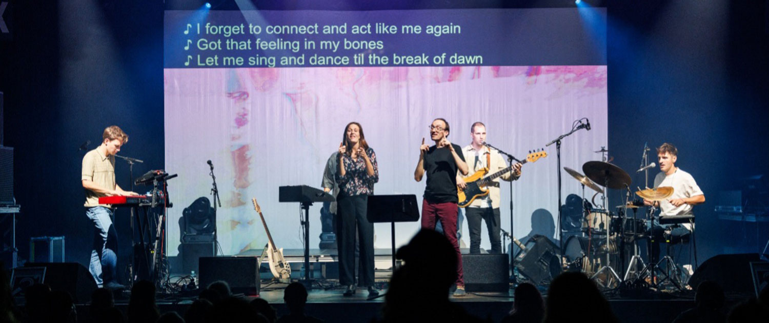 Five people are visible on a stage. They stand in front of a screen. At the top of the screen are three lines of the English lyrics to the song being sung. These lyrics are produced by sign language interpreters. The two people in the middle of the stage are sign language poets. They perform the text in sign language. © Beat the Silence with link: Das WUK | Beat the Silence
