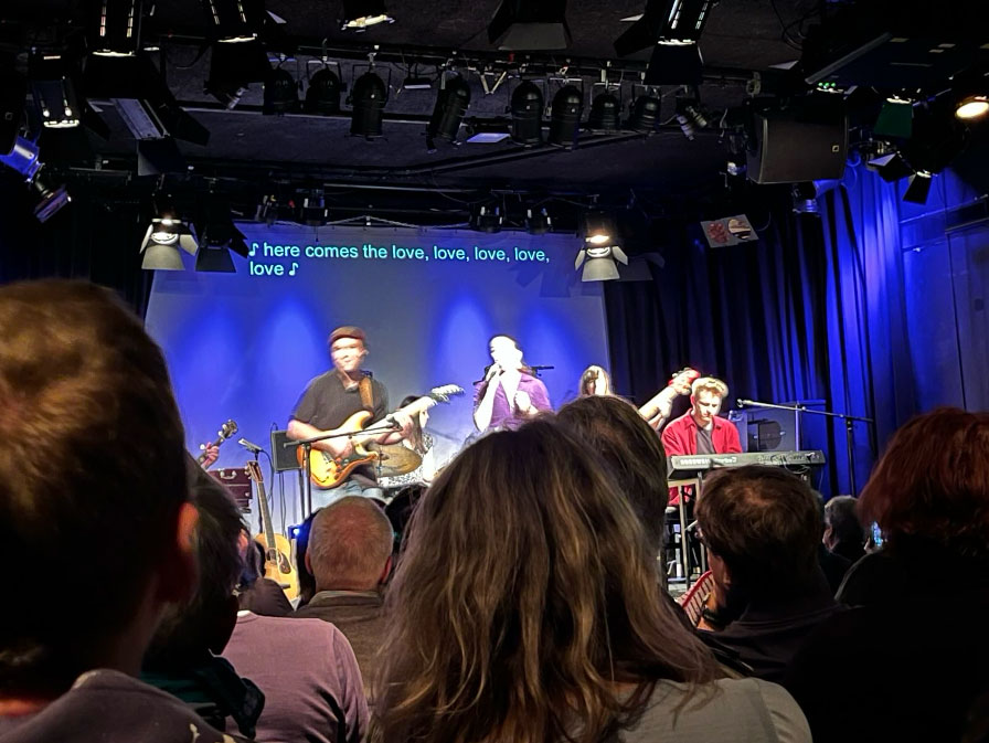 A band performing in front of a screen. At the top of the screen, two lines of the English lyrics of the song are displayed, after being produced by speech-to-text interpreters. © Beat the Silence 