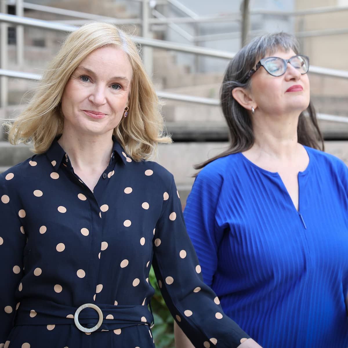 Two women in close-up. They are sitting side by side. The woman in the foreground is smiling at the camera. She is blonde and wearing a blue top with beige polka dots. It is Judith Platter. The woman in the background has her eyes closed and appears very relaxed. She has long, brown hair. She is wearing distinctive glasses with brown frames and a blue top. It is Ursula Nasswetter.