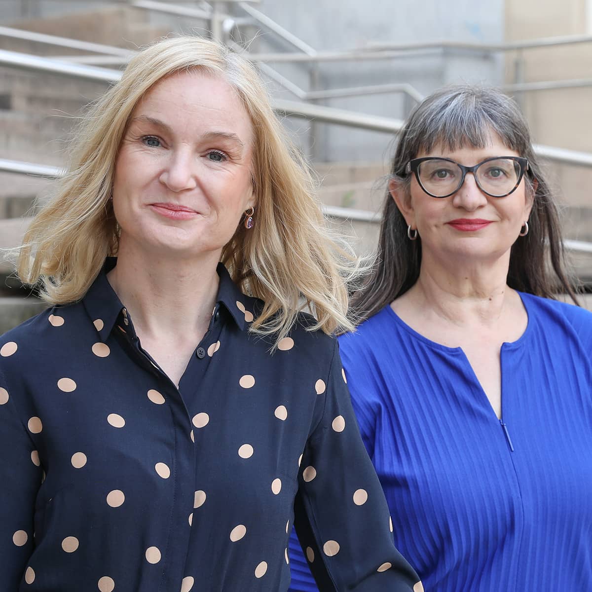 Two women in close-up. They sit side by side and smile at the camera. The woman in the foreground is blonde and wears a blue top with beige polka dots. It is Judith Platter. The woman in the background has long, brown hair. She wears distinctive glasses with brown frames and a blue top. It is Ursula Nasswetter.