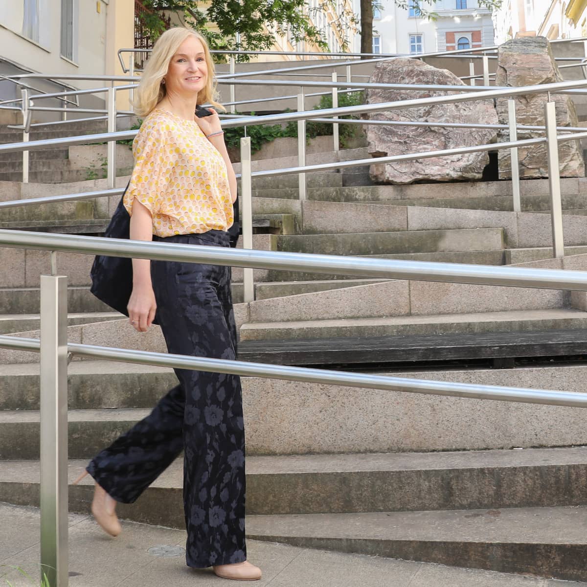 A middle-aged woman wearing a blue pantsuit with beige polka dots stands in the middle of a staircase. She smiles at the camera and leans against the metal railing of a ramp that runs up the stairs. It is Judith Platter. The staircase is the Thurnstiege, with accessible ramps in Vienna's 9th district.