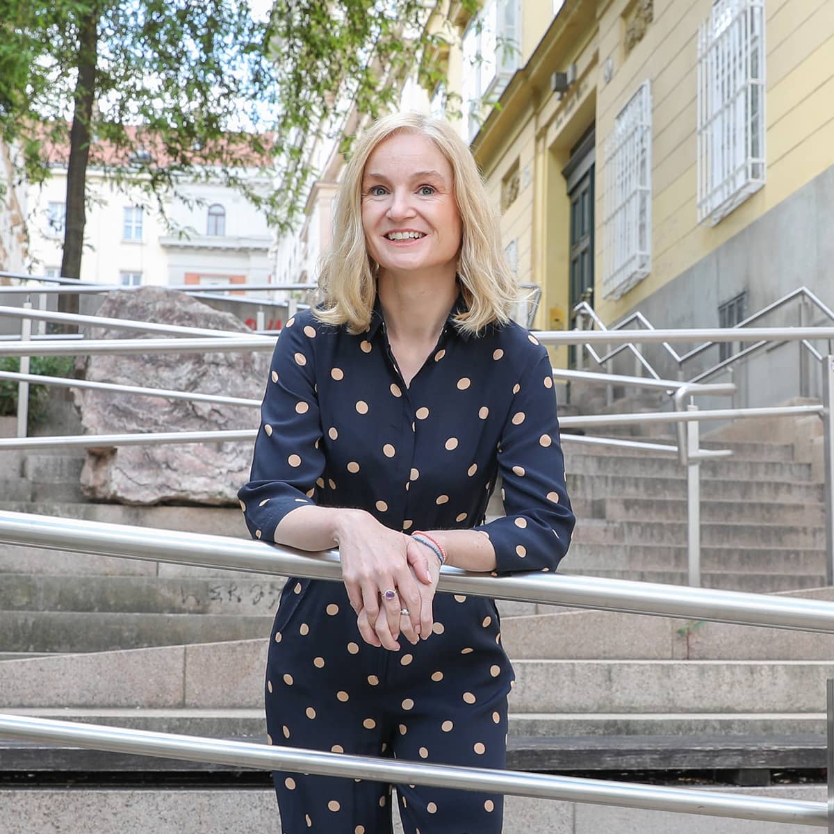A middle-aged woman wearing a blue pantsuit with beige polka dots stands in the middle of a staircase. She smiles at the camera and leans against the metal railing of a ramp that runs up the stairs. It is Judith Platter. The staircase is the Thurnstiege, with accessible ramps in Vienna's 9th district.