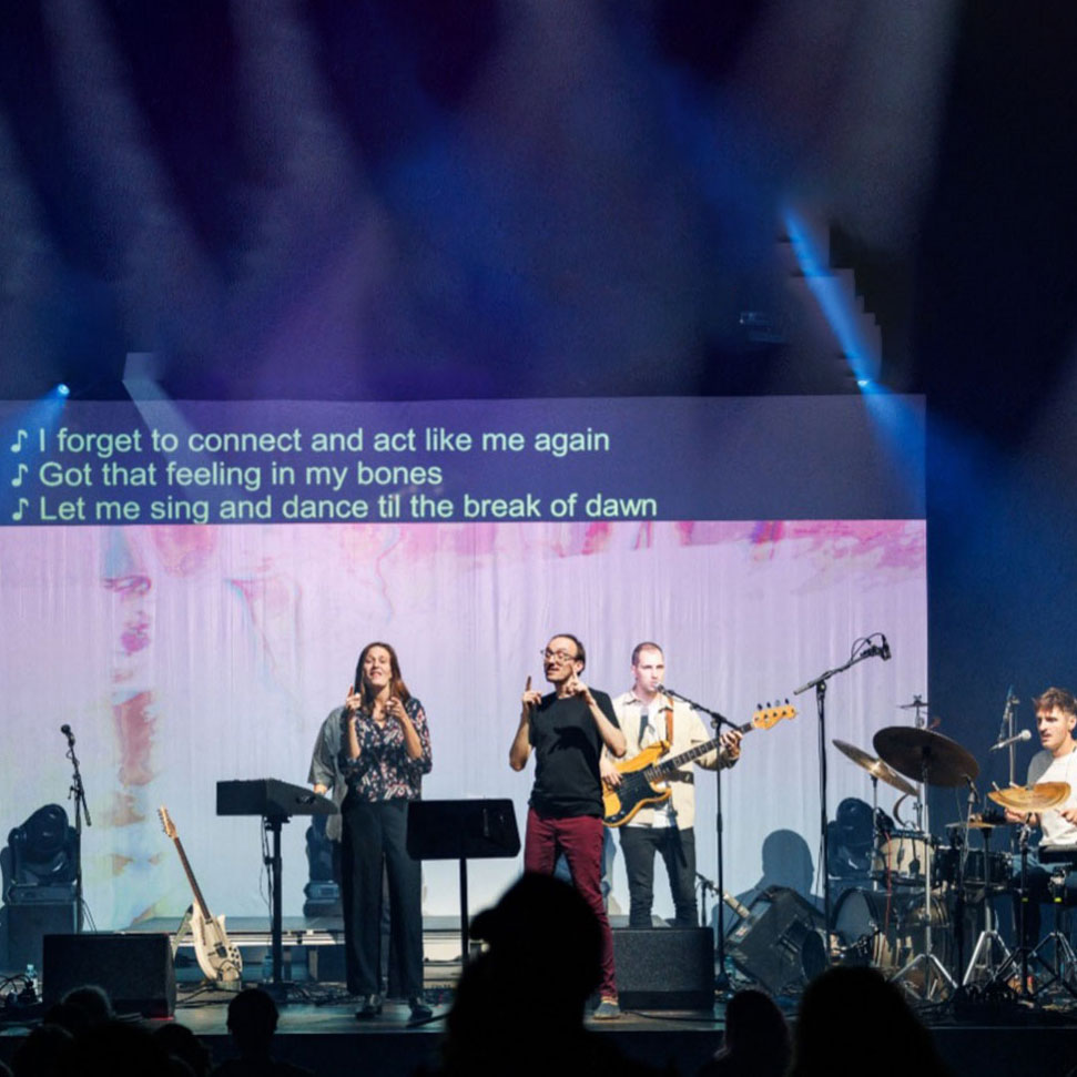 Five people on a stage standing and performing in front of a screen. At the top of the screen, three lines of the English lyrics of the song are displayed, after being produced by speech-to-text interpreters. Two people in the middle of the stage are sign language poets. They perform the text in sign language. © Beat the Silence with link: Das WUK | Beat the Silence