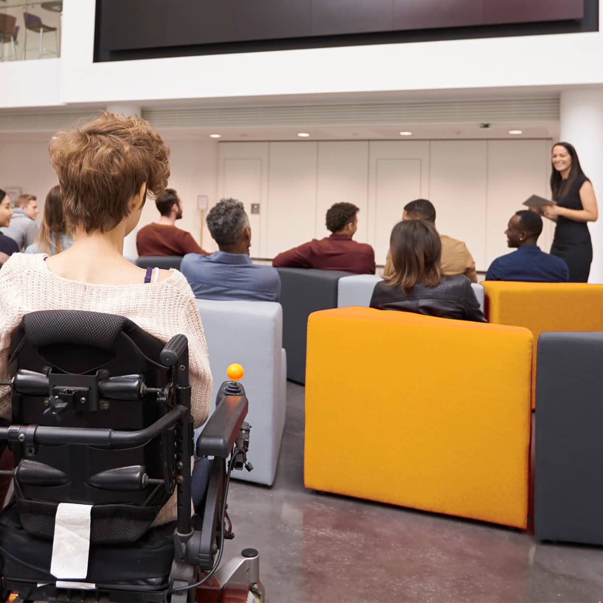 A young female speaker in an elegant black dress stands facing the audience in front of four rows of comfortable, upholstered armchairs. Around a dozen people are seated in the armchairs, they are displayed from behind. In the last row, in the center of the image, a person in a wheelchair is amongst them.