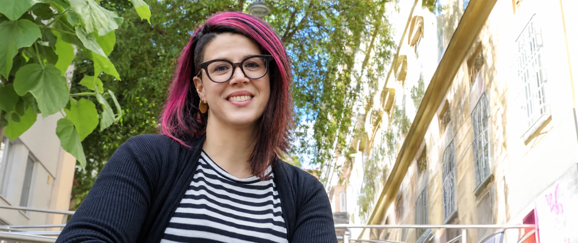 A middle-aged woman wearing a black and white sweater smiles at the camera. Her hair is pink and black, she wears round glasses. It is Verena Brinda.